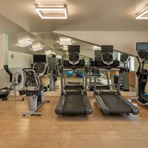 A fitness room with exercise machines, including treadmills and a rowing machine, mirrored walls, and bright overhead lighting.