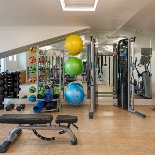 A gym with exercise equipment: weights, stability balls, mats, and machines in a mirrored room with a bench and cardio equipment.