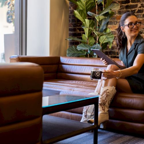 A person in glasses sits on a brown couch with a tablet and a drink, wearing a dark outfit and white cowboy boots, smiling.