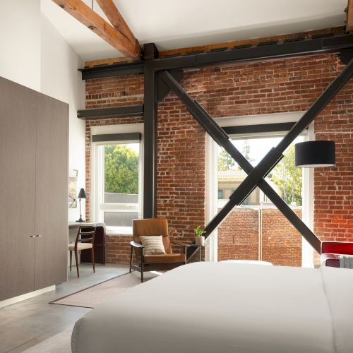 A modern loft-style room with exposed brick, wooden beams, a bed, TV, desk, and chair, blending rustic and contemporary elements seamlessly.
