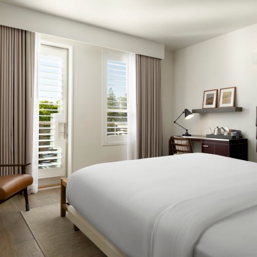 A cozy hotel room with a neatly made bed, a desk, a chair, a TV, art on the wall, and natural light coming from a window.