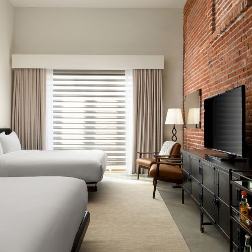 A modern hotel room with two beds, a brick wall, large TV, armchair, lamp, and a minibar cart with bottles and a plant.