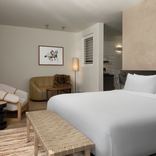 A modern, cozy bedroom with a double bed, wicker bench, lounge chairs, a wall-mounted TV, and ambient lighting.