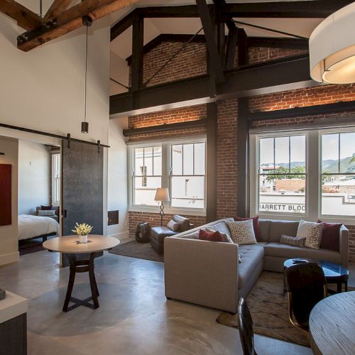 A modern living space with brick walls, exposed beams, a gray sectional sofa, wall art, and sliding door near a bedroom area.