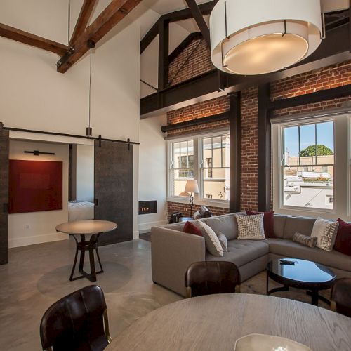 A modern loft with exposed beams, brick walls, large windows, a sectional sofa, dining table, and sliding barn doors leading to a bedroom.