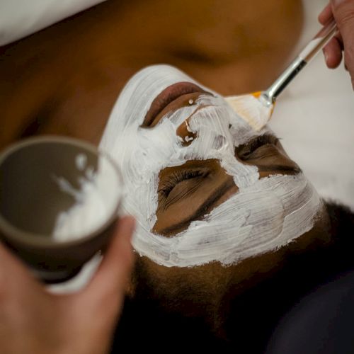A person receiving a facial treatment with a brush applying a white mask on their face.