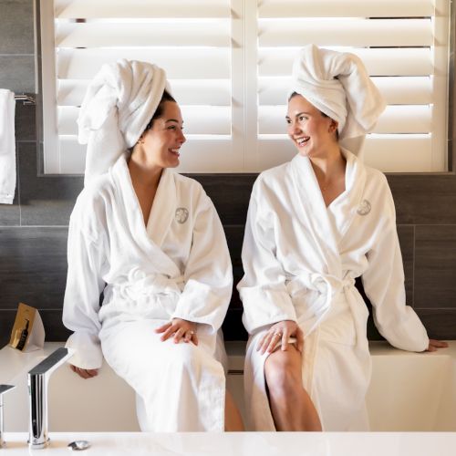 Two people in bathrobes with towels on heads, sitting on a bathtub, smiling at each other, in a modern bathroom setting.