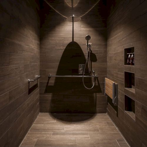The image shows a modern shower with brown tiles, a rainfall showerhead, grab bars, a foldable wooden seat, and inset shelves holding toiletries.