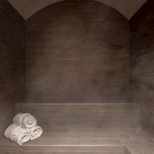 A dimly lit steam room with tiled walls, bench seating, and three rolled towels placed on the bench.