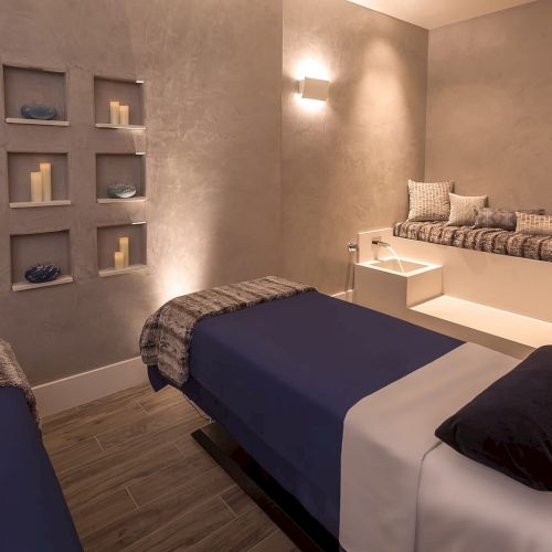The image shows a spa room with massage tables, cozy blankets, candles, and ambient lighting on a wall shelf.