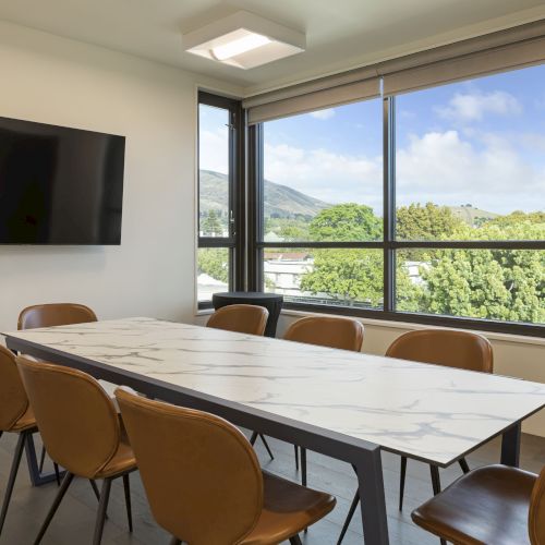 A conference room with a table, several chairs, a wall-mounted TV, and large windows with a view of trees and hills outside.