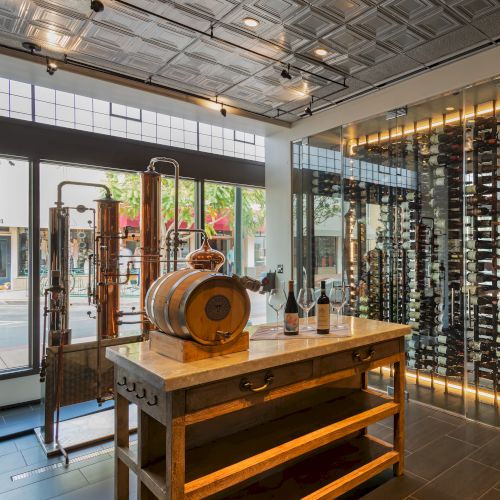 The image shows a modern wine display with bottles on glass shelves, a wooden table, a small barrel, and distillation equipment.