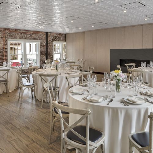 Elegant dining room set for an event; round tables with white tablecloths, plates, glasses, utensils, and chairs are arranged neatly.