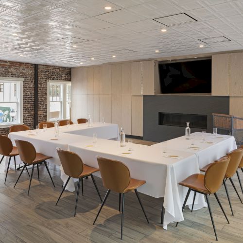 A conference room with a U-shaped table setup, brown chairs, white tablecloths, water bottles, and a large screen on the wall.