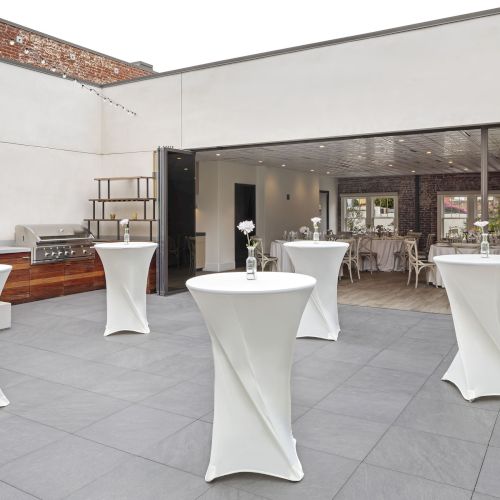 Outdoor patio with tall tables covered in white cloths, small flowers in vases, and a built-in grill. Indoor dining area visible.