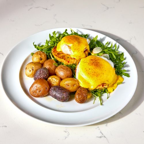 The image shows eggs Benedict on a bed of greens with roasted potatoes on a white plate.