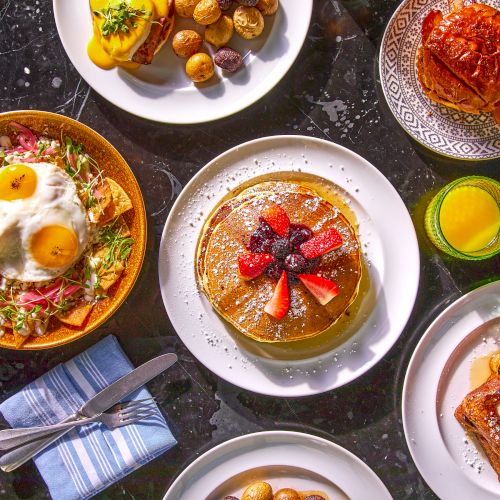 The image shows a table with pancakes, a breakfast bowl with eggs, toast with fruit, a croissant, potatoes, orange juice, and napkins.