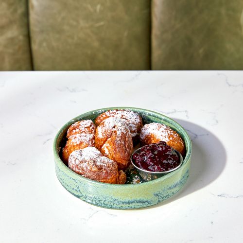 A bowl of pastries dusted with powdered sugar sits on a table, with a small dish of berry jam beside them.