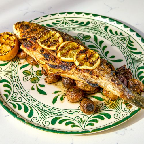 A grilled fish garnished with lemon slices on a decorative plate with roasted vegetables.