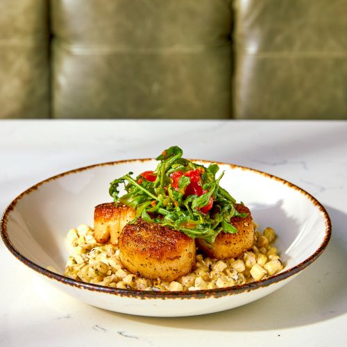 The image shows seared scallops on a bed of creamy corn, topped with greens and diced tomatoes, served in a white bowl.