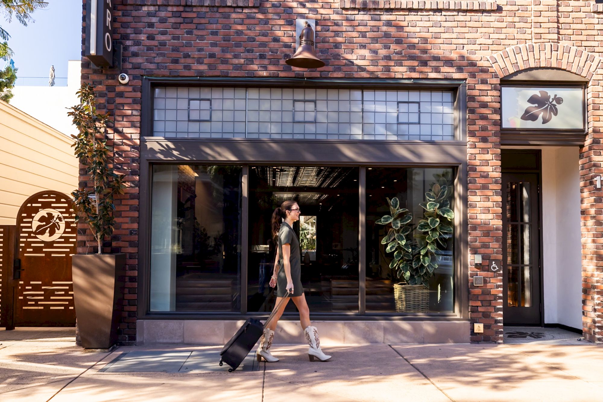 A person walks with a suitcase in front of a brick building with large windows and plants, under a sunny sky.