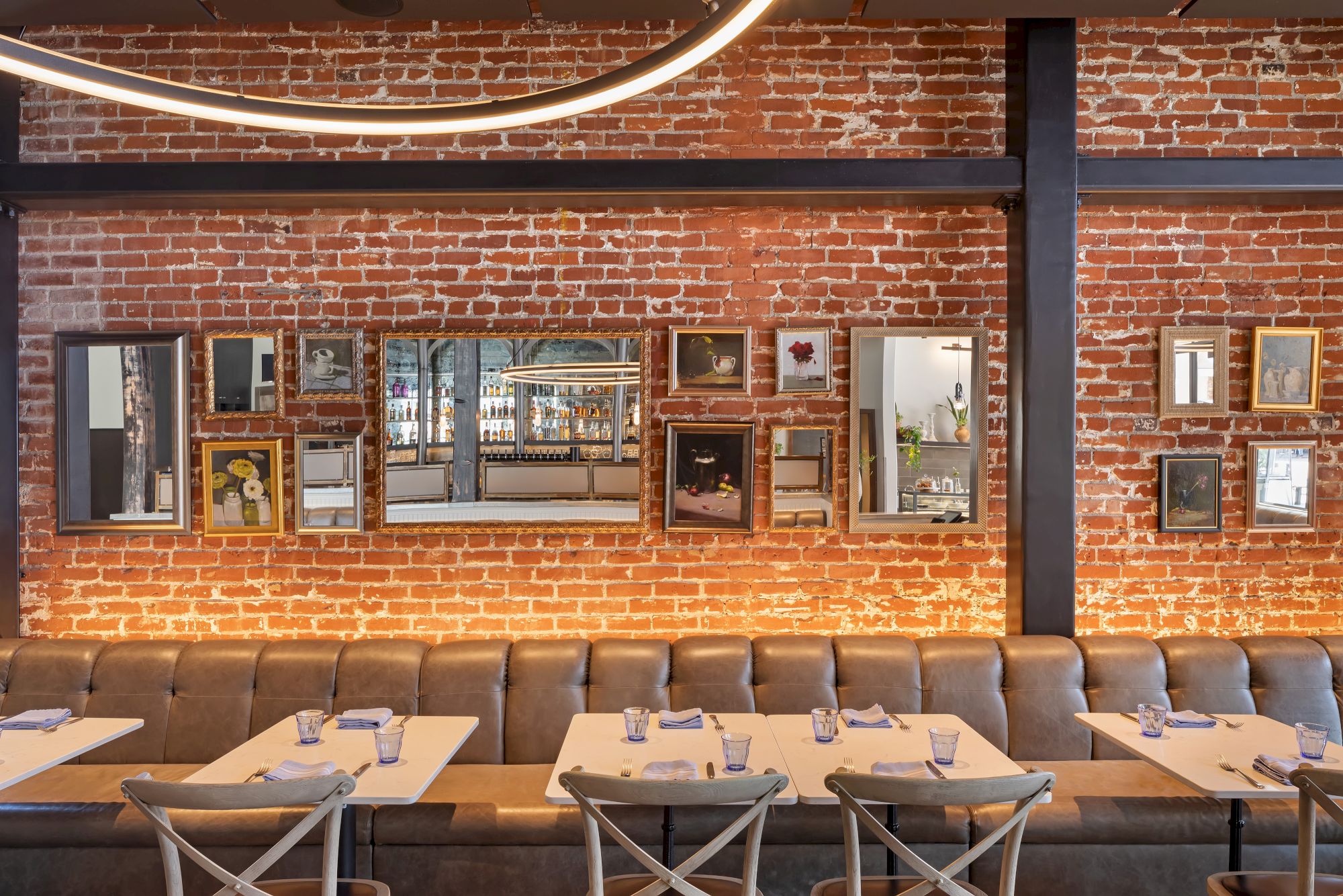 A stylish restaurant interior with brick walls, mirrors, framed art, wooden chairs, and neatly set tables for dining.