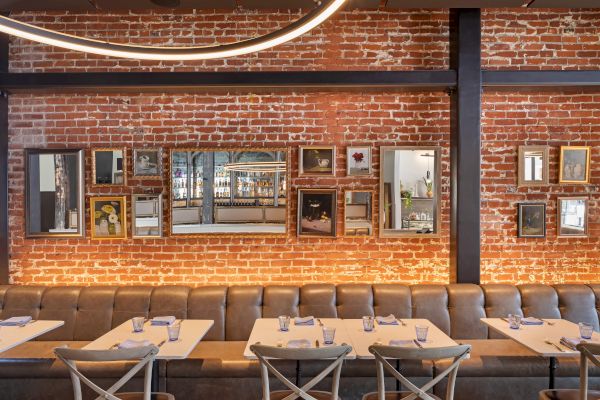 A restaurant with a brick wall, mirrors, and framed art. Tables set with glasses, napkins, and utensils line the wall with a leather bench.