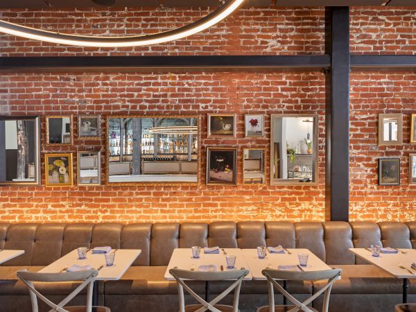 A restaurant with a brick wall, mirrors, and framed art. Tables set with glasses, napkins, and utensils line the wall with a leather bench.