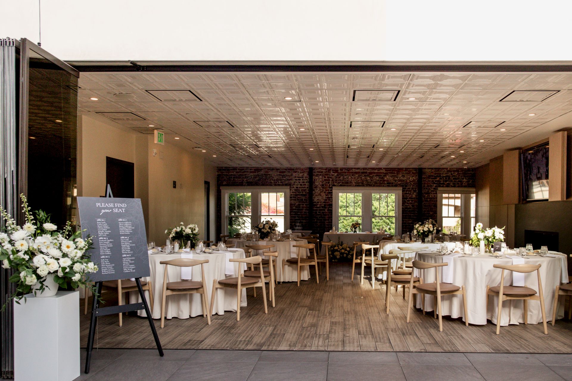 The image shows an elegant banquet hall with round tables, chairs, floral arrangements, and a menu board by the entrance.