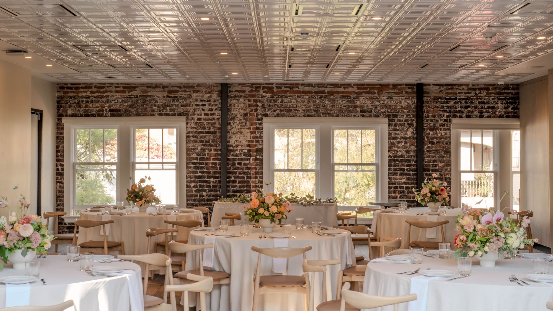 The image shows an elegantly decorated room with round tables, covered in white cloth, and floral centerpieces, set for an event.