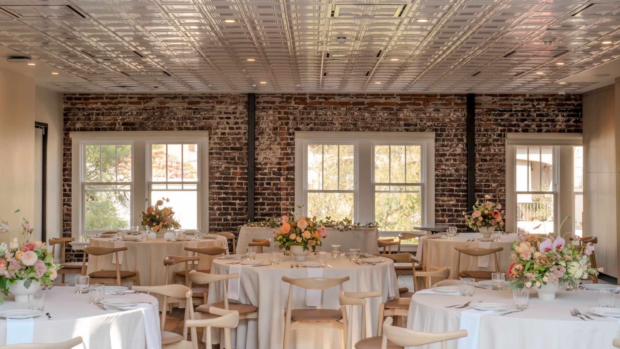 The image shows an elegantly decorated room with round tables, covered in white cloth, and floral centerpieces, set for an event.