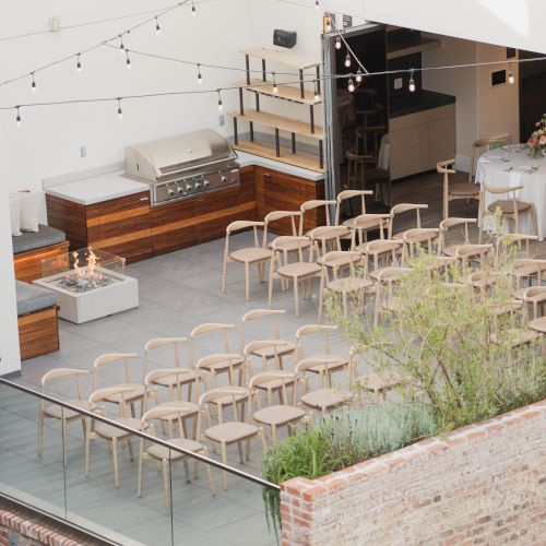 The image shows an outdoor seating area with wooden chairs arranged in rows, surrounded by brick walls and string lights overhead.