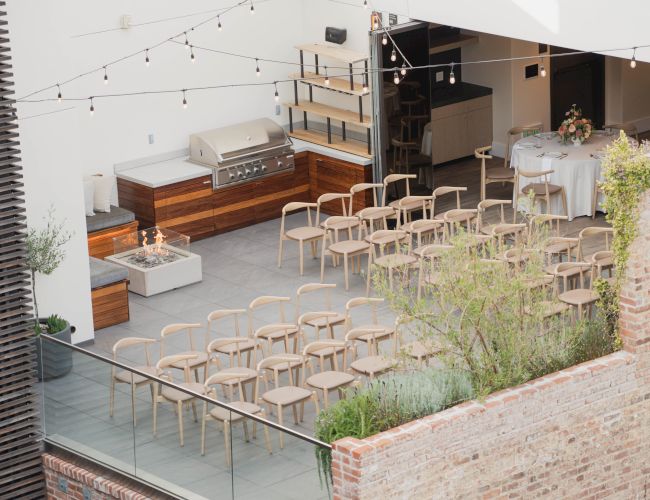 The image shows an outdoor seating area with wooden chairs arranged in rows, surrounded by brick walls and string lights overhead.