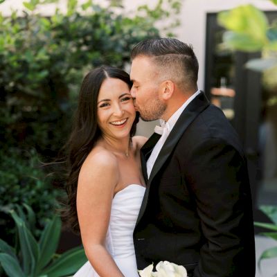 A couple is dressed in wedding attire; the man kisses the woman's cheek in a garden setting. They appear to be very happy together.