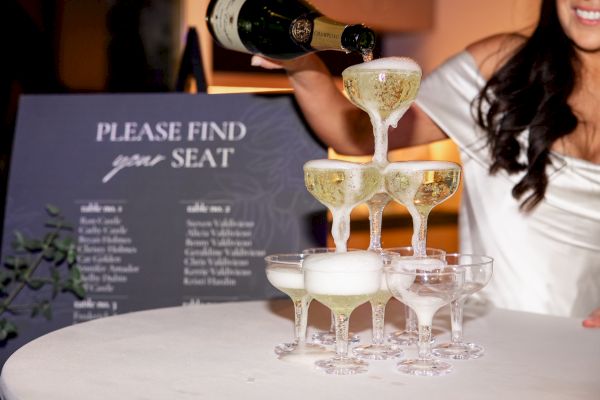 A champagne tower is being poured, with a seating chart in the background showing guest names at an event.