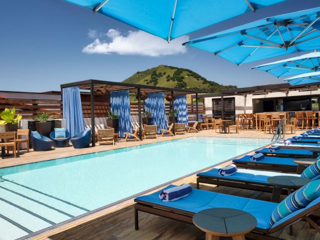 The image shows a stylish outdoor pool area with blue lounge chairs and umbrellas, surrounded by a scenic mountain view in the background.
