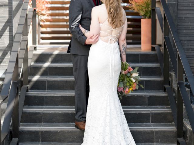 A couple is standing on stairs; the woman in a white dress holds flowers, and they embrace near a modern entrance.