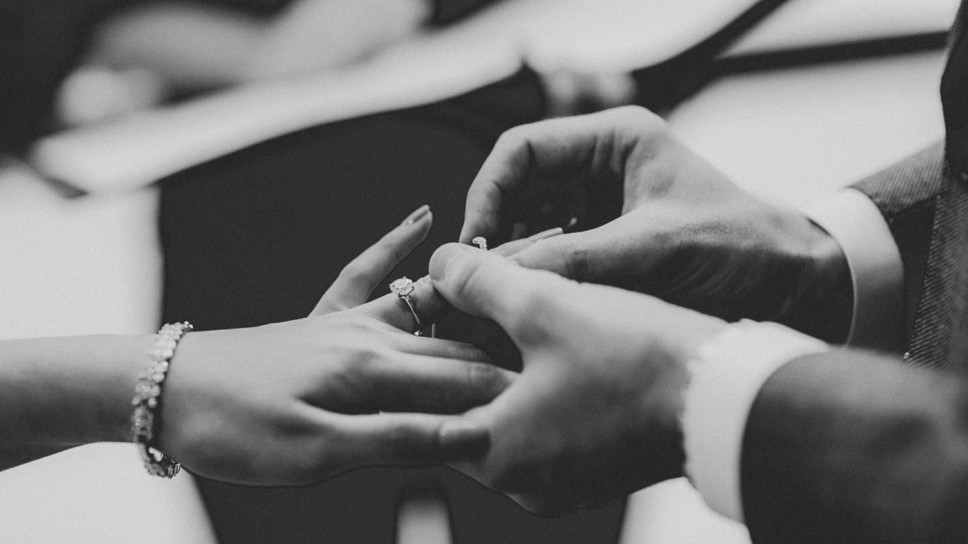 A black and white image of a person placing a ring on another's finger, symbolizing a wedding or engagement ceremony.