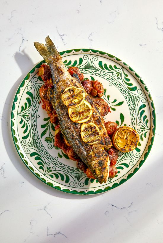 A whole grilled fish garnished with lemon slices, served on a decorative plate with seasoned potatoes or vegetables.