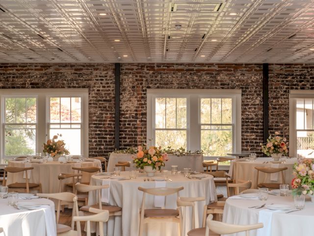 The image shows a decorated dining room with round tables, flowers, brick walls, and a patterned ceiling, set for an event.