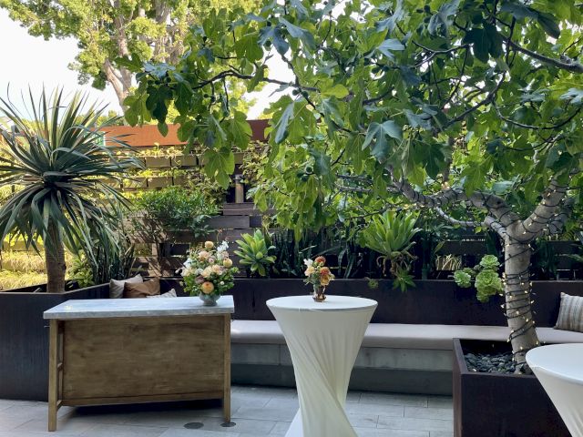 An outdoor patio with plants, a tree, a small table with a white cover, and a wooden table adorned with flower arrangements.