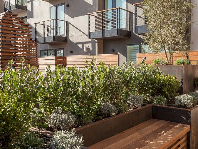 The image shows a modern apartment building with balconies, surrounded by landscaped greenery and a wooden bench in the foreground.