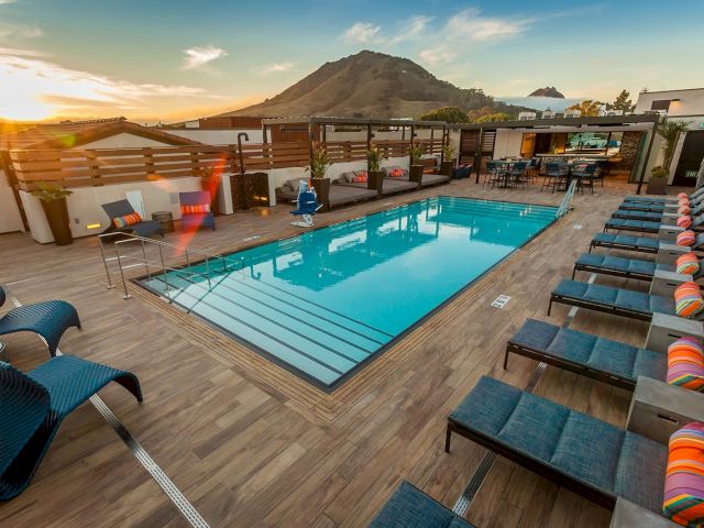 A rooftop pool with lounge chairs, colorful pillows, and mountain views at sunset. The setting is stylish and inviting for relaxation.