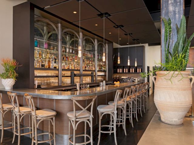 A stylish bar features wooden chairs, a well-stocked backlit shelf, pendant lights, and large decorative plants.