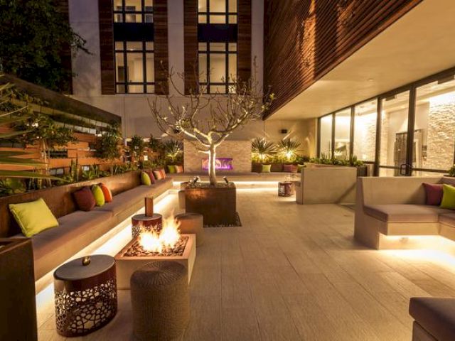 A modern outdoor patio with cozy seating, a fire pit, decorative lighting, pillows, and plants creating a warm and inviting atmosphere.