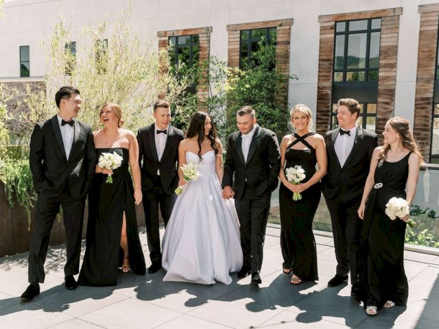 A wedding party poses outdoors; the bride in a white gown, bridesmaids in black dresses, and groomsmen in black suits.