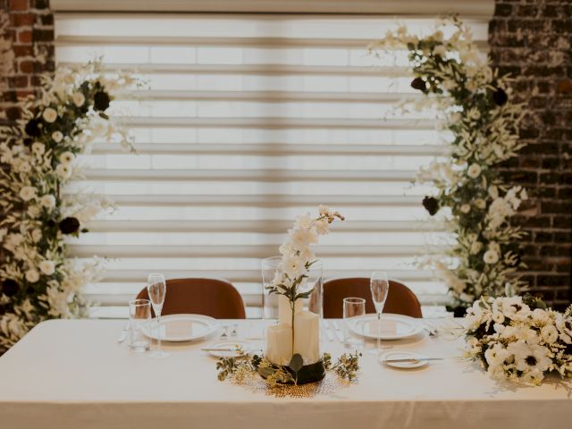 A decorated table with elegant floral arrangements, plates, and wine glasses set against a backdrop of brick and large window blinds.