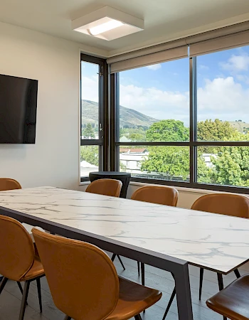 A modern conference room with a long marble table, tan chairs, a wall-mounted TV, and large windows showing a green landscape and blue sky.