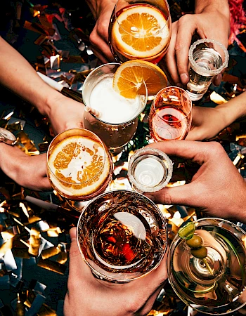 A circle of hands cheers with colorful cocktails and sliced oranges, confetti glitters above&mdash;celebration and friends to toast together.