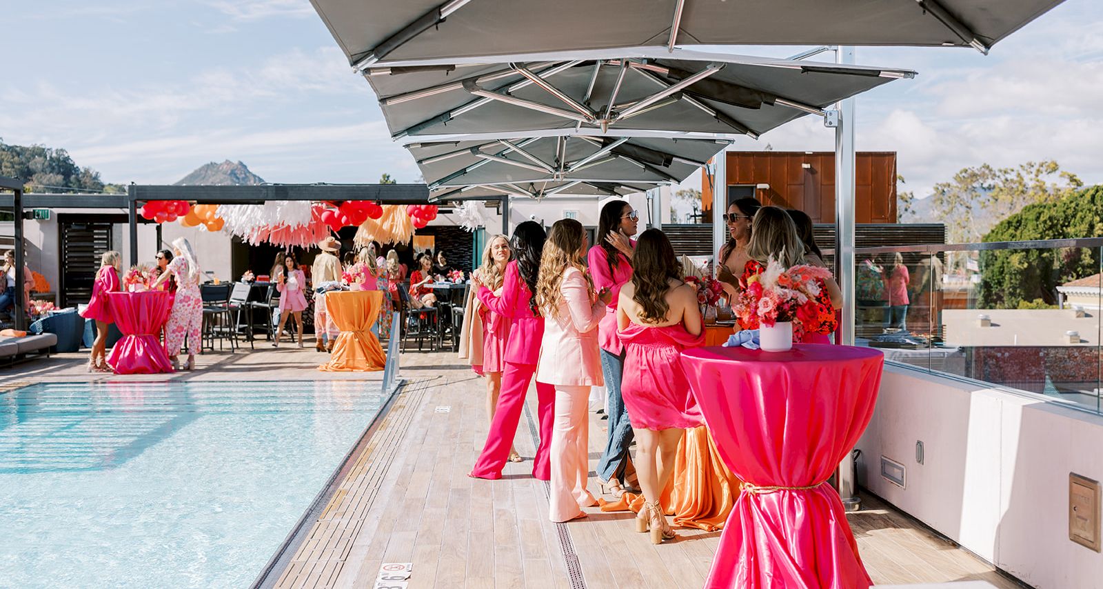People gather by a poolside bar and lounge under large umbrellas, dressed in bright colors for a stylish outdoor party.